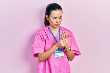 Young brunette woman wearing doctor uniform and stethoscope suffering pain on hands and fingers,...