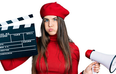 Young brunette teenager holding video film clapboard and megaphone puffing cheeks with funny face....