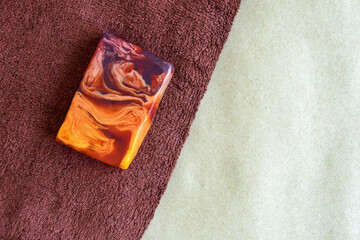 Piece of orange-brown natural handmade soap on braun fluffy terry towel. Hygiene, body care and health concept. Top view, flat lay. Copy space. Selective focus.