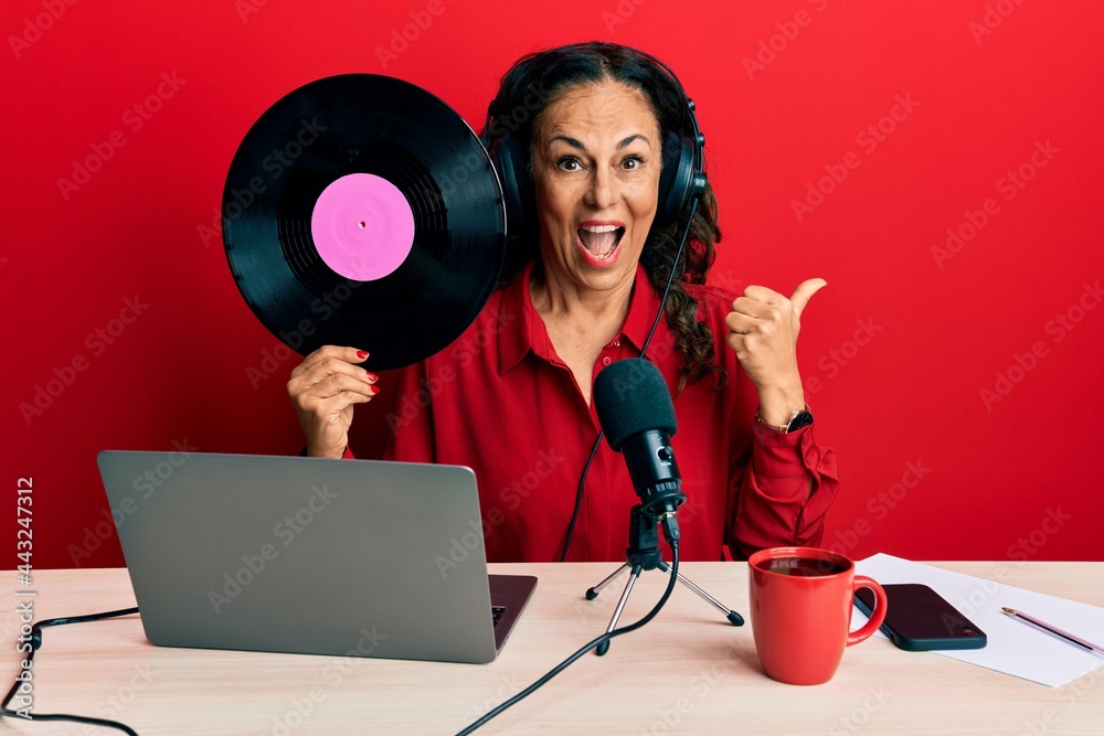 Poster beautiful middle age woman working at radio studio holding vinyl disc pointing thumb up to the side 