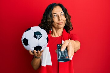 Middle age hispanic woman football hooligan holding ball using tv control smiling looking to the side and staring away thinking.