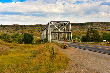 Two lane highway heading across steel bridge over small river into beautiful badlands country.