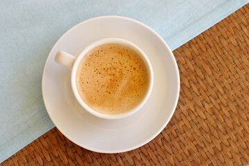 Cup of cappuccino coffee in a white saucer on wooden table with a blue tablecloth, top view
