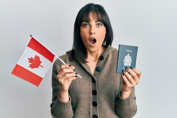 Naklejka premium Young hispanic woman holding canada flag and passport afraid and shocked with surprise and amazed expression, fear and excited face.