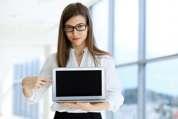 Beautiful female specialist with laptop computer standing in modern office and smiling charmingly. Working on design, data analysis, plan strategy. Business people concept