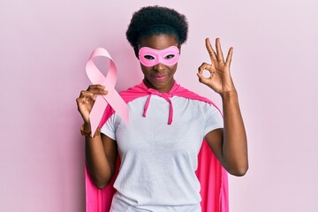 Young african american girl wearing superhero costume holding pink cancer ribbon doing ok sign with fingers, smiling friendly gesturing excellent symbol