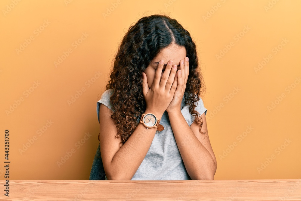 Wall mural teenager hispanic girl wearing casual clothes sitting on the table with sad expression covering face