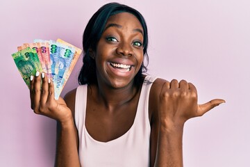 Young african american woman holding south african rand banknotes pointing thumb up to the side...