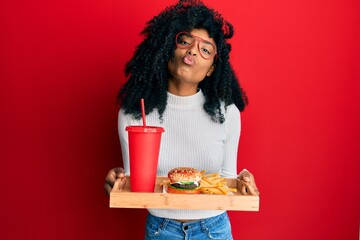 African american woman with afro hair eating a tasty classic burger with fries and soda looking at...