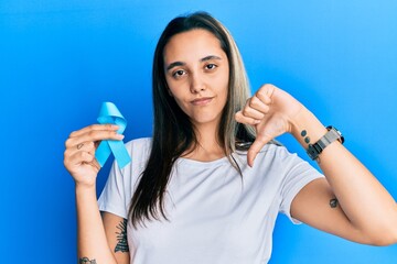 Young hispanic woman holding blue ribbon with angry face, negative sign showing dislike with thumbs down, rejection concept