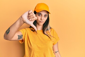 Young hispanic woman wearing delivery uniform and cap looking unhappy and angry showing rejection and negative with thumbs down gesture. bad expression.