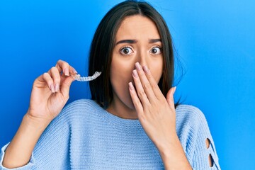 Young hispanic girl holding invisible aligner orthodontic covering mouth with hand, shocked and...