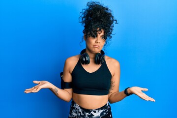 Young hispanic woman wearing gym clothes and using headphones clueless and confused expression with...