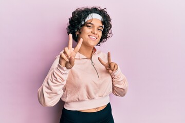 Young hispanic woman with curly hair wearing sportswear smiling looking to the camera showing fingers doing victory sign. number two.