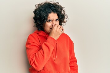 Fototapeta na wymiar Young hispanic woman with curly hair wearing casual sweatshirt smelling something stinky and disgusting, intolerable smell, holding breath with fingers on nose. bad smell
