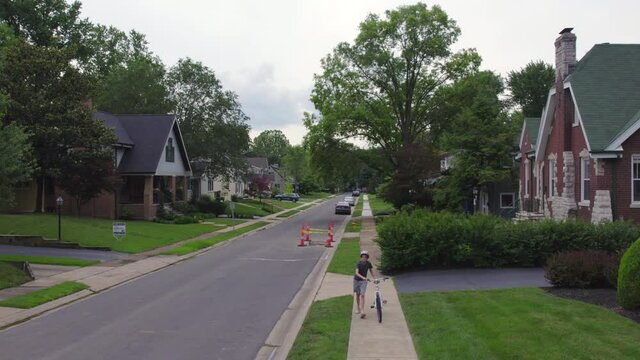 Boy walks his bike up the sidewalk towards camera as a car drives past the other way.