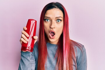 Young caucasian woman holding shampoo bottle scared and amazed with open mouth for surprise,...