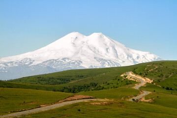 Elbrus is the highest peak in Russia and Europe. Snow mountain on the background of green fields and blue sky. Kabardino-Balkaria, view of Elbrus from Jila-Su.