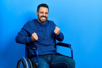 Handsome hispanic man with beard sitting on wheelchair very happy and excited doing winner gesture with arms raised, smiling and screaming for success. celebration concept.