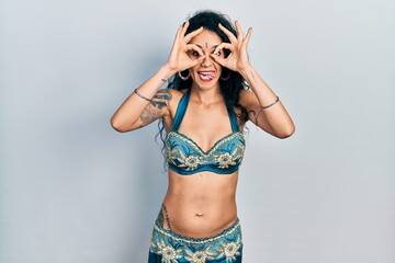 Young woman wearing bindi and traditional belly dance clothes doing ok gesture like binoculars sticking tongue out, eyes looking through fingers. crazy expression.