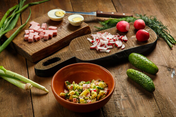 Okroshka in a clay plate on a table on a wooden table next to an egg sausage and cucumbers on planks.