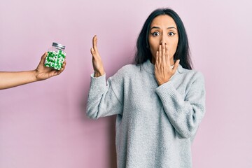 Young hispanic girl rejecting jar of pills with hand covering mouth with hand, shocked and afraid...
