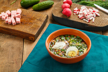 Okroshka in a clay plate on the table on a tablecloth next to vegetables on boards.
