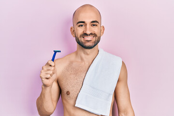 Young bald man shirtless holding razor looking positive and happy standing and smiling with a confident smile showing teeth - obrazy, fototapety, plakaty