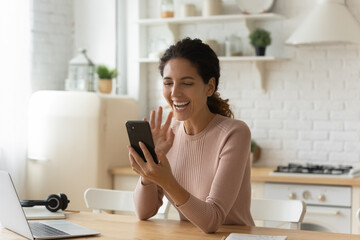 Joyful young hispanic caucasian woman looking at cellphone screen, enjoying distant web camera call using mobile video call zoom application, involved in pleasant distant conversation in kitchen.
