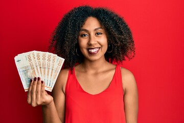 Beautiful african american woman with afro hair holding 100 danish krone banknotes looking positive and happy standing and smiling with a confident smile showing teeth