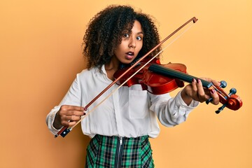 Beautiful african american woman with afro hair playing classical violin afraid and shocked with surprise and amazed expression, fear and excited face.