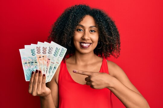 Beautiful african american woman with afro hair holding czech koruna banknotes smiling happy pointing with hand and finger