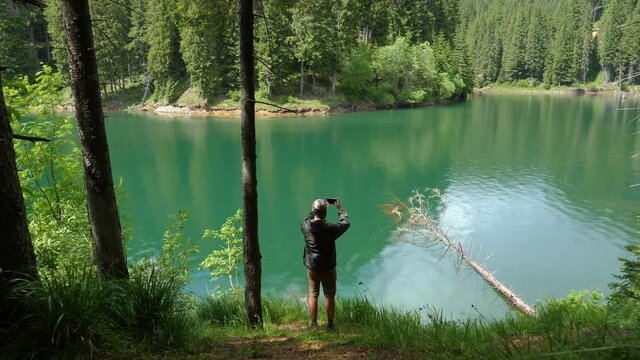 Man taking photos through the forest on the shore of the mountain lake. Walking in the woods on the edge of a turquoise mountain lake. Mountain forest lake. Young boy dressed in camouflage hiking.