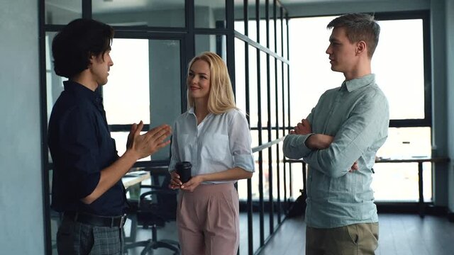 Side view of three positive young business people having fun conversation during coffee break in modern office. Happy coworkers enjoy pleasant communication, discuss project on background of window.