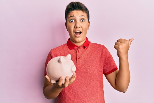 Teenager hispanic boy holding piggy bank pointing thumb up to the side smiling happy with open mouth