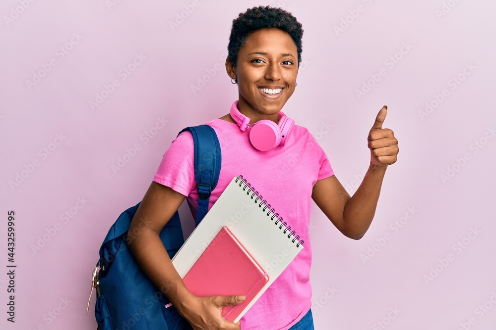 Wall mural Young african american girl wearing school bag and holding books smiling happy and positive, thumb up doing excellent and approval sign