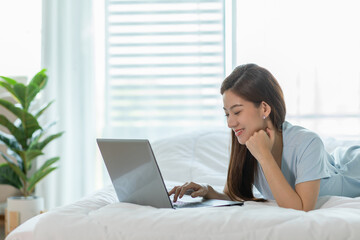 Beautiful Attractive Asian young woman lying on bed and using computer laptop for using social media and shopping online relax in cozy bedroom in holiday,Lifestyle in home Concept