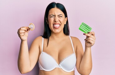 Beautiful brunette woman holding condom and birth control pills sticking tongue out happy with funny expression. - obrazy, fototapety, plakaty