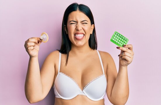 Beautiful brunette woman holding condom and birth control pills sticking tongue out happy with funny expression.
