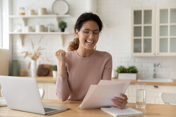 Emotional happy young woman in glasses reading paper correspondence letter with good news, celebrating getting excellent test results, last mortgage payment notification, feeling overjoyed in kitchen.
