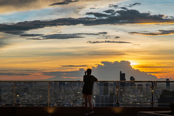 Obraz premium Bangkok, Thailand - 02 Jul 2021 : A young woman relaxing on rooftop terrace with overlooking the city at sunset view. Night city, Blurred town on background, Copy space, Selective focus.