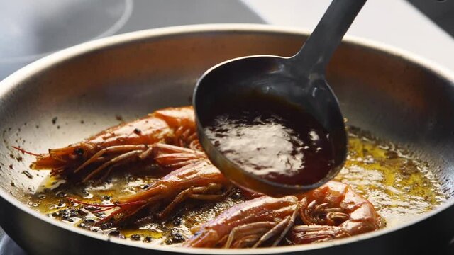 Cook pours shrimps on a grill with sauce from ladle. Closeup of king prawns frying in a pan. Chef frying royal shrimps on a hot pan. Macro sea food preparation.