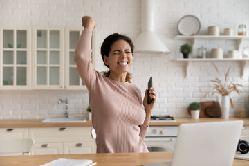 Overjoyed millennial sincere woman making yes gesture, holding smartphone in hands, celebrating...