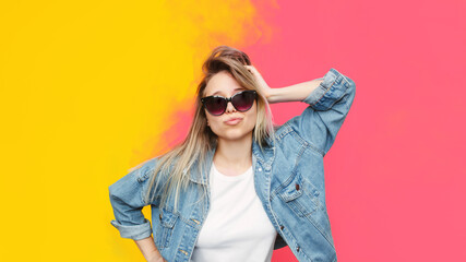 A young pretty caucasian stylish blonde woman in a white t-shirt, blue denim jeans jacket, black sunglasses poses with her hand on a head isolated on a color yellow and pink background.Beauty, fashion