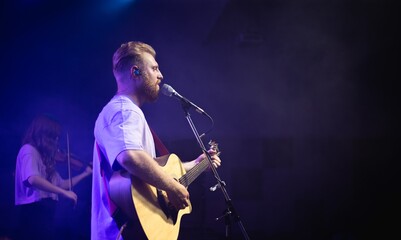 A young man with a beard holds an acoustic guitar in his hand and sings into a microphone stands on the stage