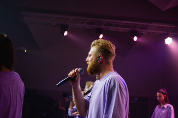 A young man with a beard holds an acoustic guitar in his hand and sings into a microphone stands on...
