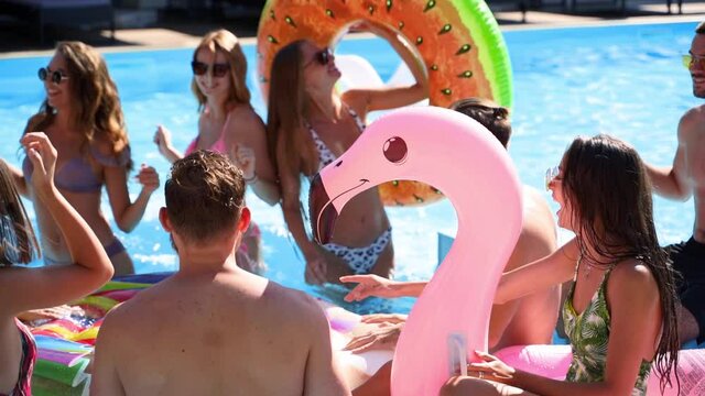 Friends have party in a private villa swimming pool. Happy young people in swimwear dancing, bonding and clubbing with floaties and inflatable mattress in luxury resort on sunny day. Slow motion.