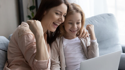 We did it. Excited mother and little daughter embrace on cozy couch hold laptop on knees show...