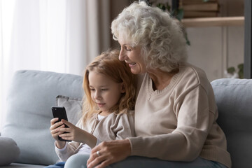 Using cell with grandma. Preteen girl grandkid cuddle on comfy sofa with older grandmother show granny funny photo on phone. Aged hoary grandparent enjoy online mobile game with beloved grandchild