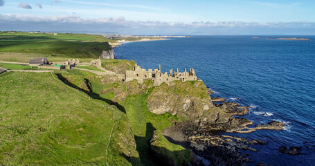 Ruins of medieval Dunluce Castle on a steep cliff near Bushmills. Northern coast of County Antrim, Northern Ireland, UK. Aerial view in sunrise light. Far view of Portrush resort in the background - obrazy, fototapety, plakaty